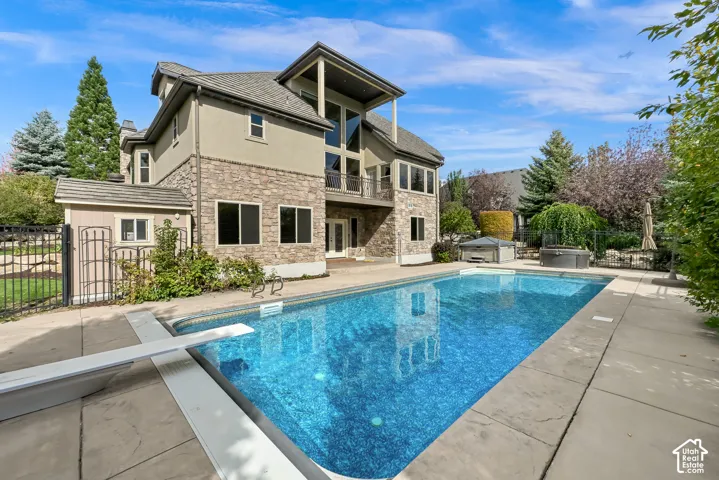 Multi-story exterior featuring stone veneer, stucco siding, a rectangular swimming pool with a diving board, an elevated balcony with metal railing, and concrete pool decking