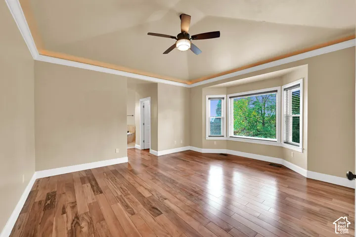 Room featuring wood-finish flooring, a tray ceiling, crown molding, a bay window, and a ceiling fan with integrated lighting