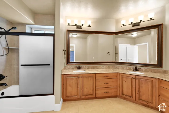 Bathroom featuring a dual-sink vanity with light wood cabinetry, stone-surfaced countertop, integrated mirrors with wood framing, and a tiled shower-tub combination with a frosted glass enclosure