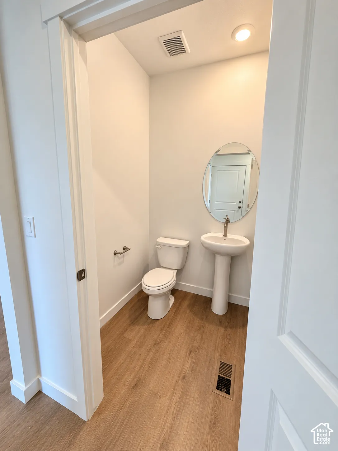 Powder room featuring wood-finish flooring, a white pedestal sink, an oval wall-mounted mirror, and a standard-height toilet