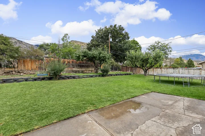 Spacious backyard featuring a concrete patio, expansive grass lawn, wood perimeter fencing, and mature trees against a mountain backdrop