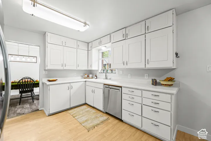 Kitchen featuring white cabinetry with shaker-style doors, light-toned wood-finish flooring, solid-surface countertops, integrated stainless steel dishwasher, and a window-mounted sink