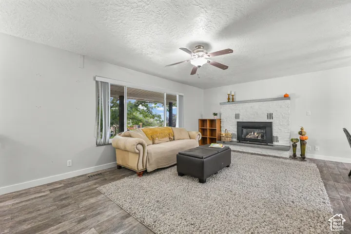 Living area featuring wood-finish flooring, white-painted textured masonry fireplace with an inset hearth, ceiling fan with integrated lighting, and large picture window