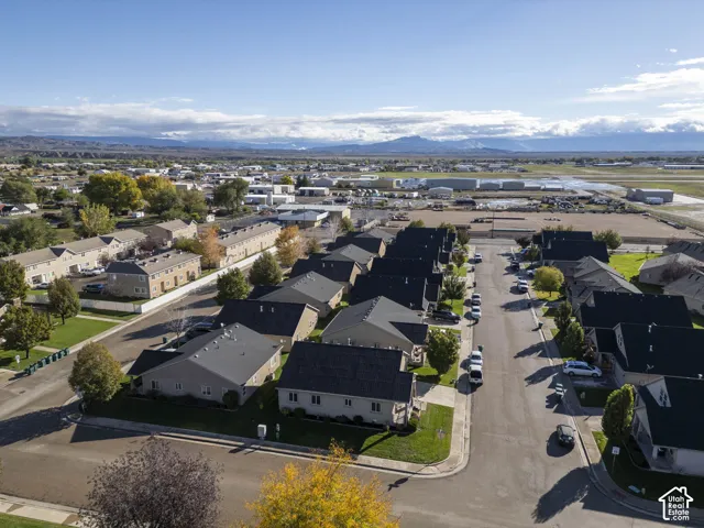 Aerial perspective of a residential neighborhood featuring paved streets, suburban housing with pitched roofing, manicured lawn areas, mature trees, and a mountain range backdrop