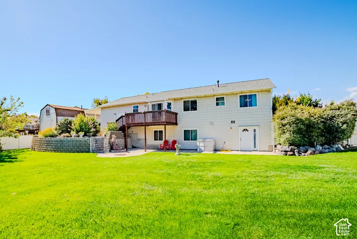 Expansive backyard featuring a large grass lawn, a raised stone retaining wall, a raised wooden deck, and a paved patio area