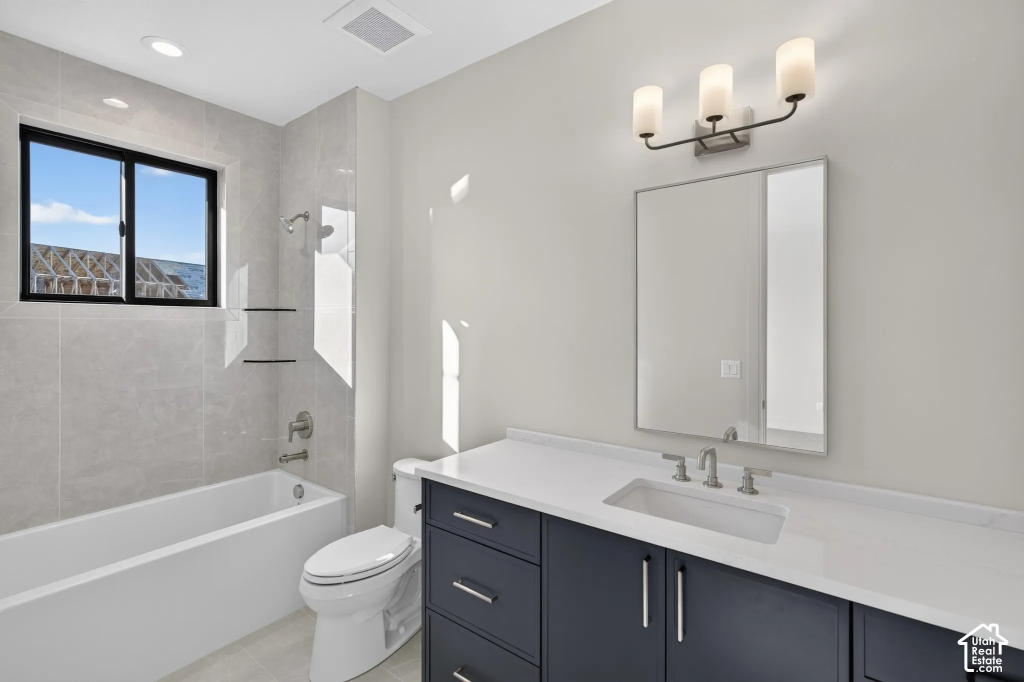 Bathroom featuring a vanity with dark cabinetry and a white countertop, an undermount sink, a framed mirror with three-light fixture, and a tile-surround tub with wall-mounted glass shelving