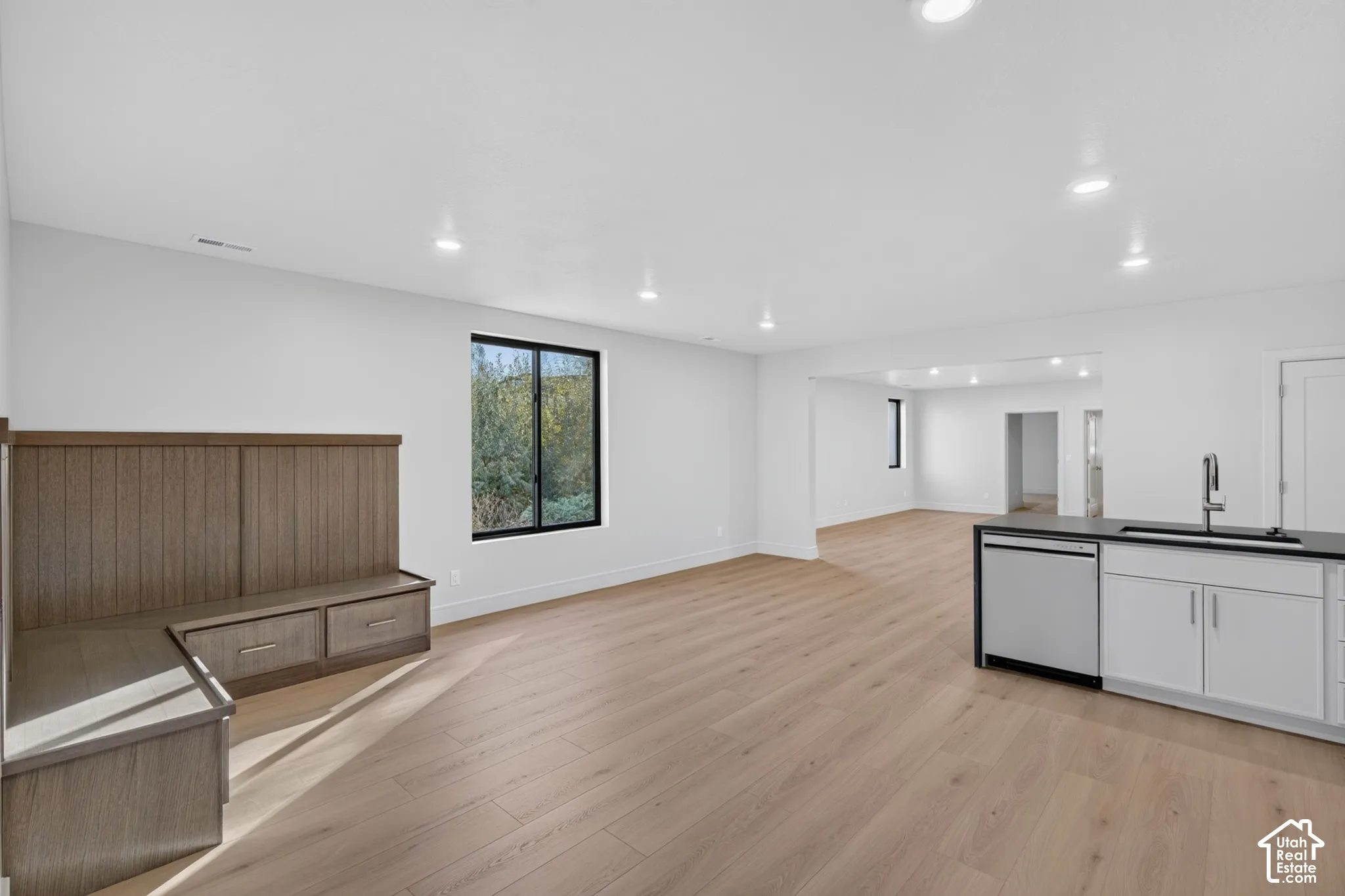 Open-concept interior featuring light wood-finish flooring, built-in wooden bench seating with storage drawers, recessed lighting, and a kitchen island with a stainless steel dishwasher and sink