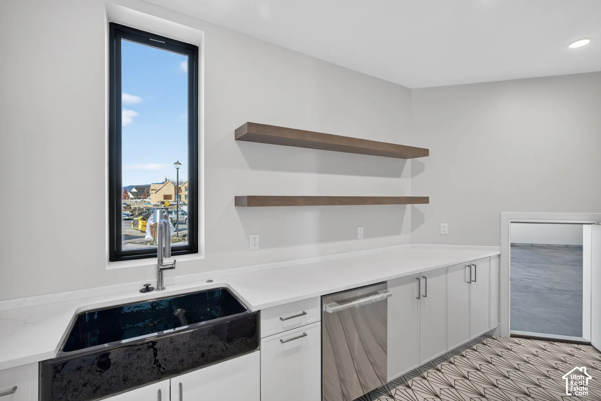 Black apron-front sink with a chrome gooseneck faucet, quartz countertops, white flat-panel cabinetry, stainless steel dishwasher, and dual wood-finish floating shelves