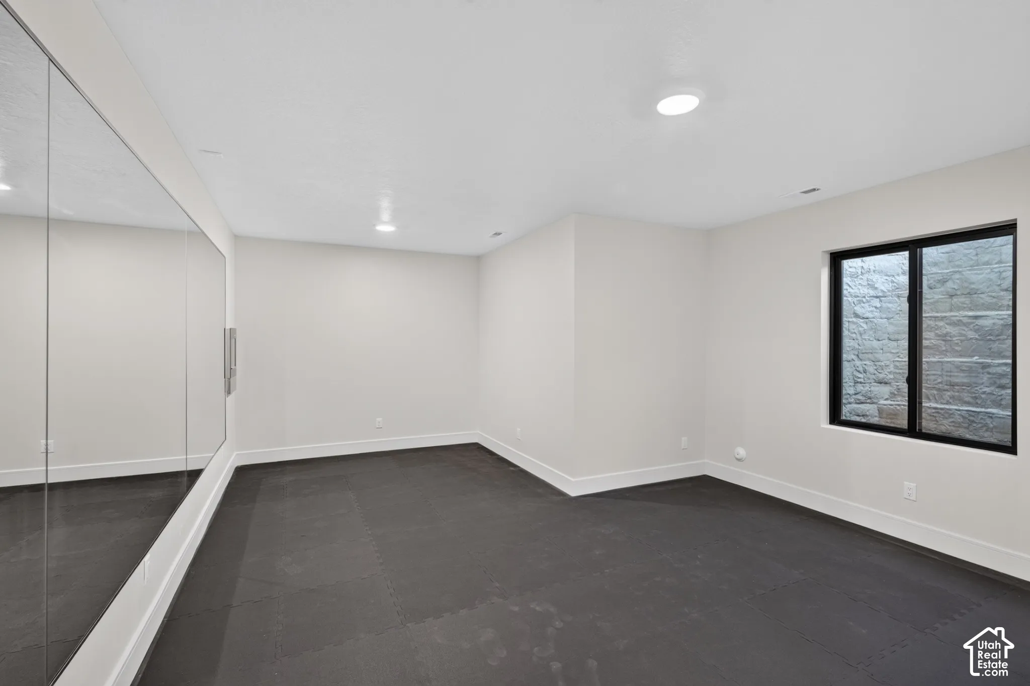 Room featuring wall-to-wall mirror, dark resilient rubberized flooring, flush-mount lighting, and a window with a stone-retaining wall well