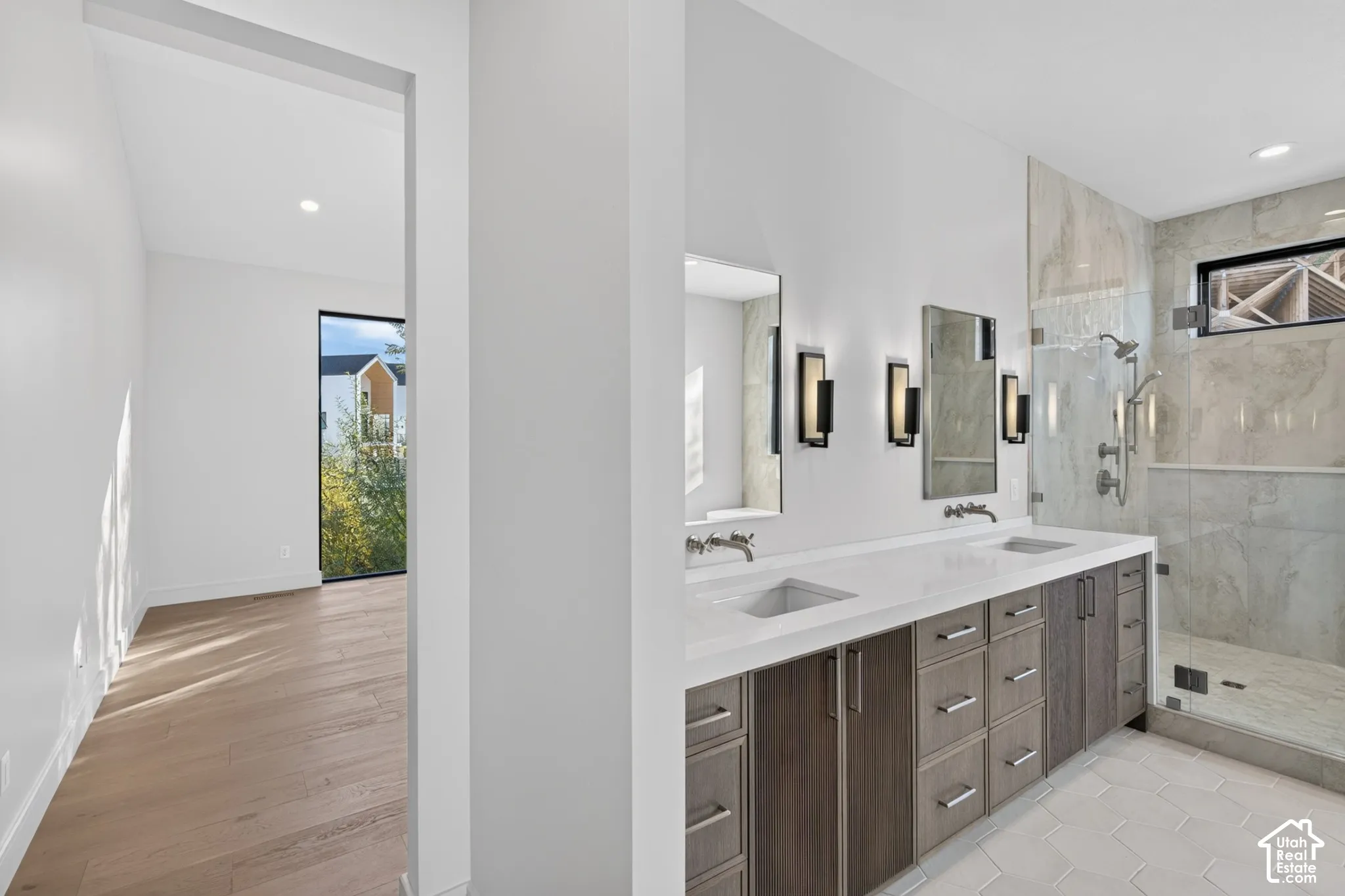 Double vanity with a composite surface, wood-finish cabinetry, wall-mounted faucets, large-format tile shower enclosure, and hexagon floor tile