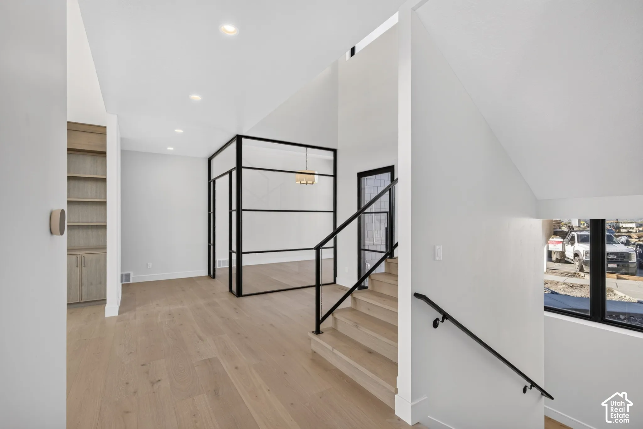 Light-toned wood-finish flooring, recessed ceiling lighting, built-in floor-to-ceiling cabinetry, black metal-framed glass partition, and staircase featuring black metal railings