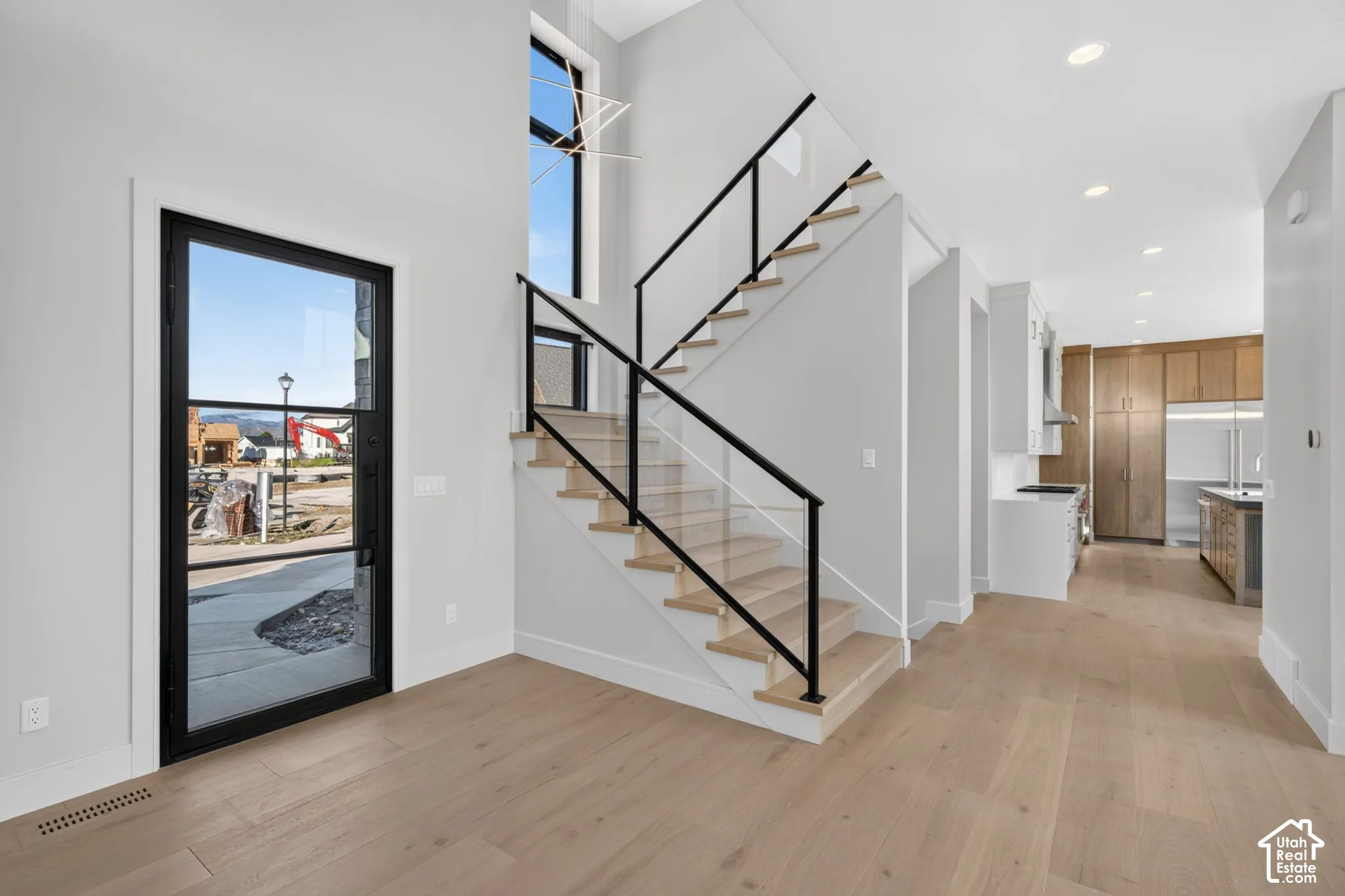 Staircase with light-toned wood treads and a matte black metal railing featuring glass panels, light-toned wood-finish flooring, recessed ceiling lighting, and a full-height glass-paneled exterior door