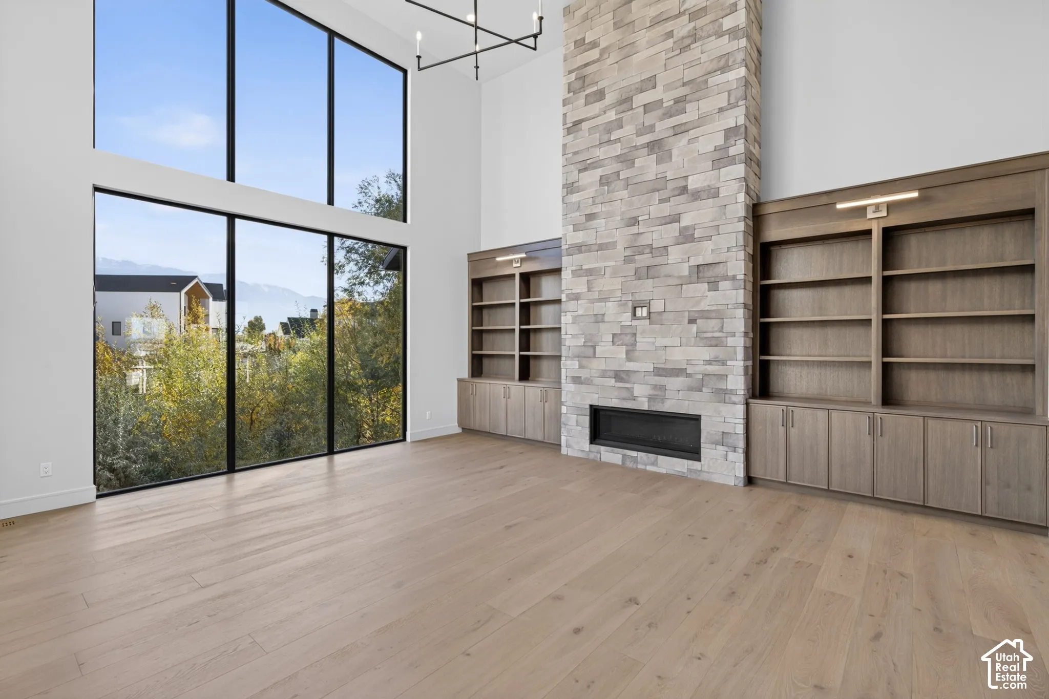 Double-height floor-to-ceiling windows, full-height stone fireplace surround, custom built-in cabinetry, wood-finish flooring, and minimalist linear chandelier
