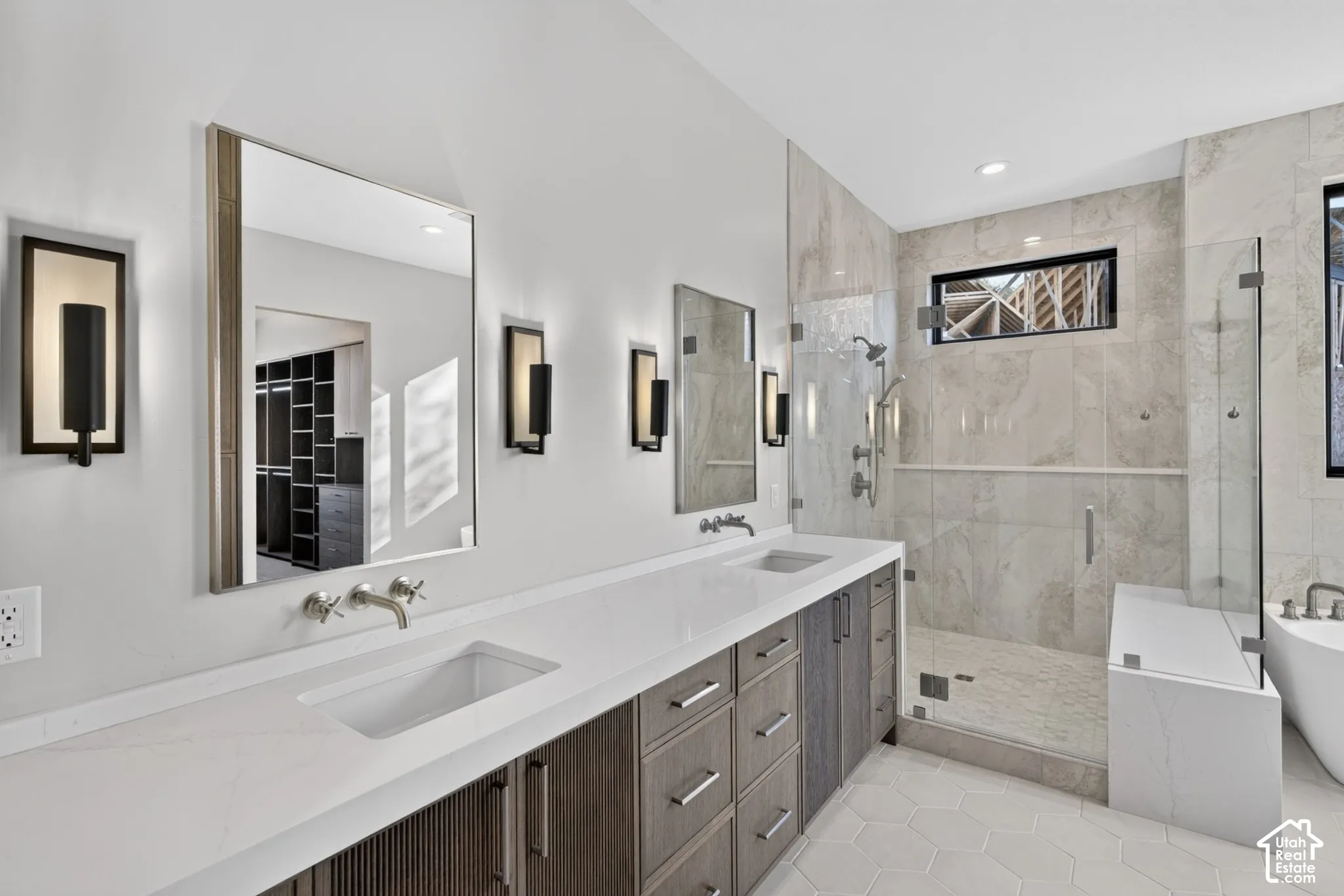 Dual-sink vanity with quartz countertop, wall-mounted faucets, glass-enclosed walk-in shower with stone-finish tile, and hexagonal floor tile