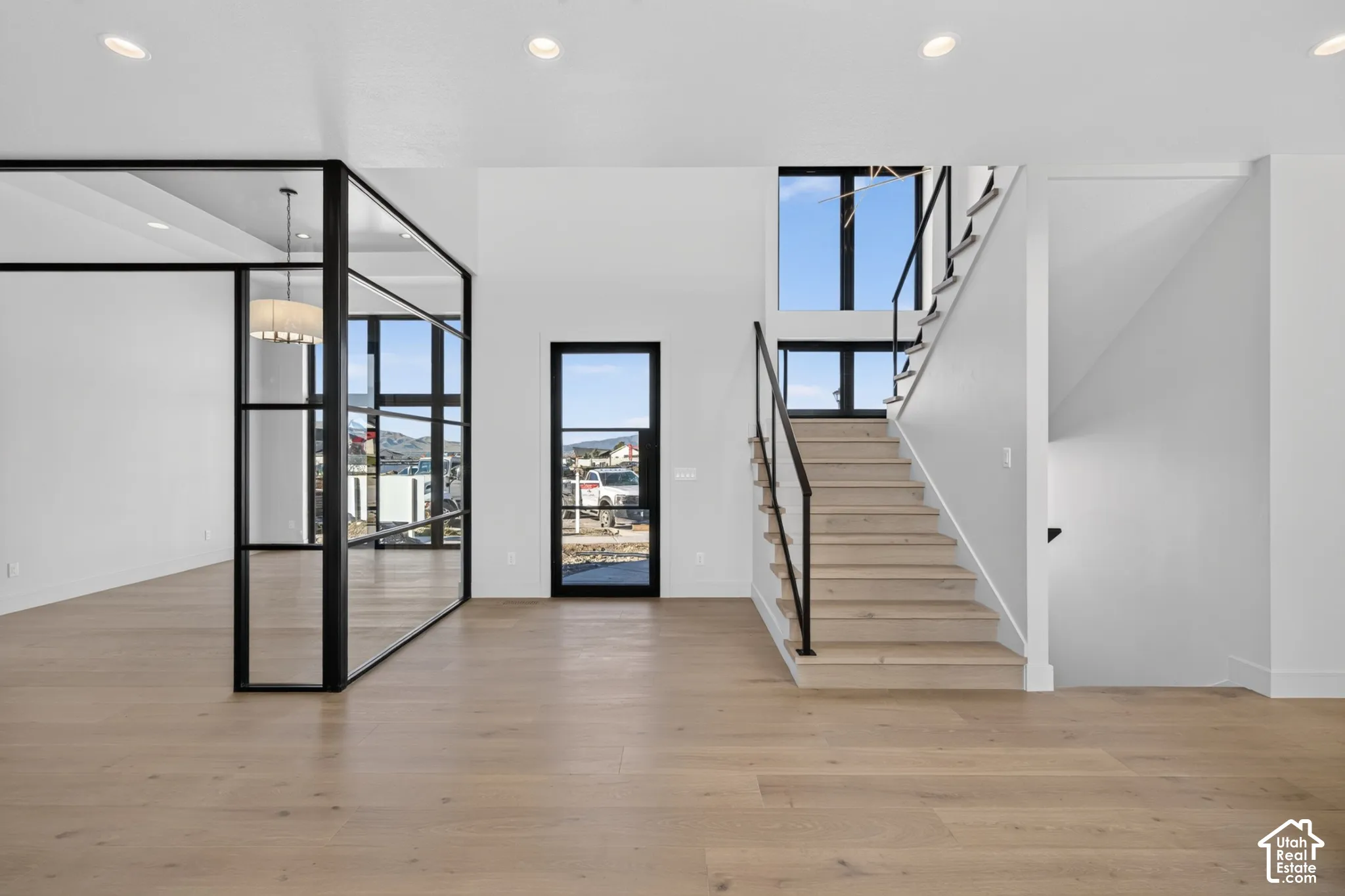 Open-concept interior featuring light-toned wood-finish flooring, a floor-to-ceiling glass-enclosed partition with black metal framing, a statement staircase with wooden treads and a black metal railing, and high-reaching windows