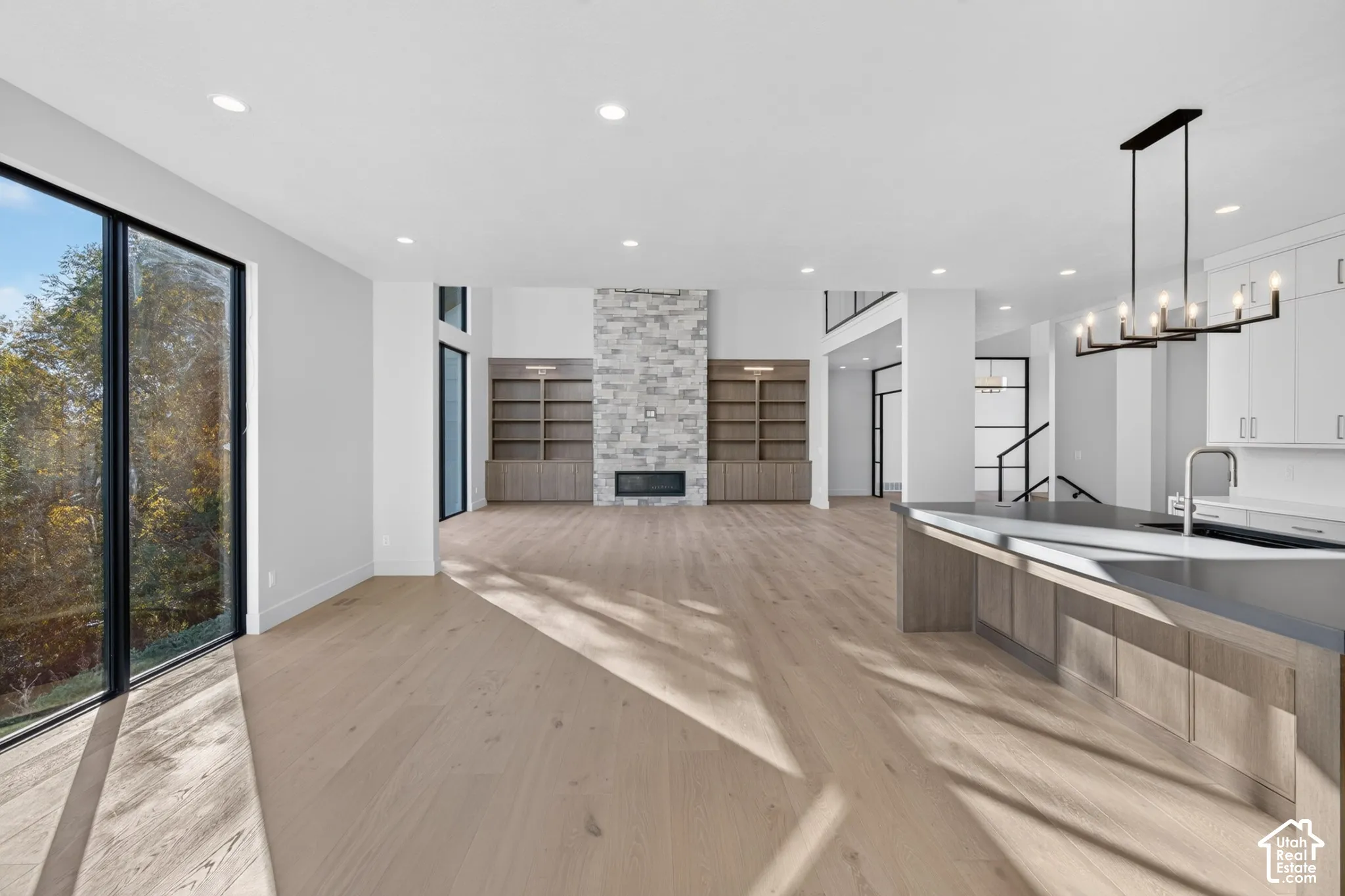 Open-concept living space featuring wood-finish flooring, a full-height stone fireplace, custom built-in cabinetry, floor-to-ceiling glass panel windows, and a central kitchen island with modern pendant lighting