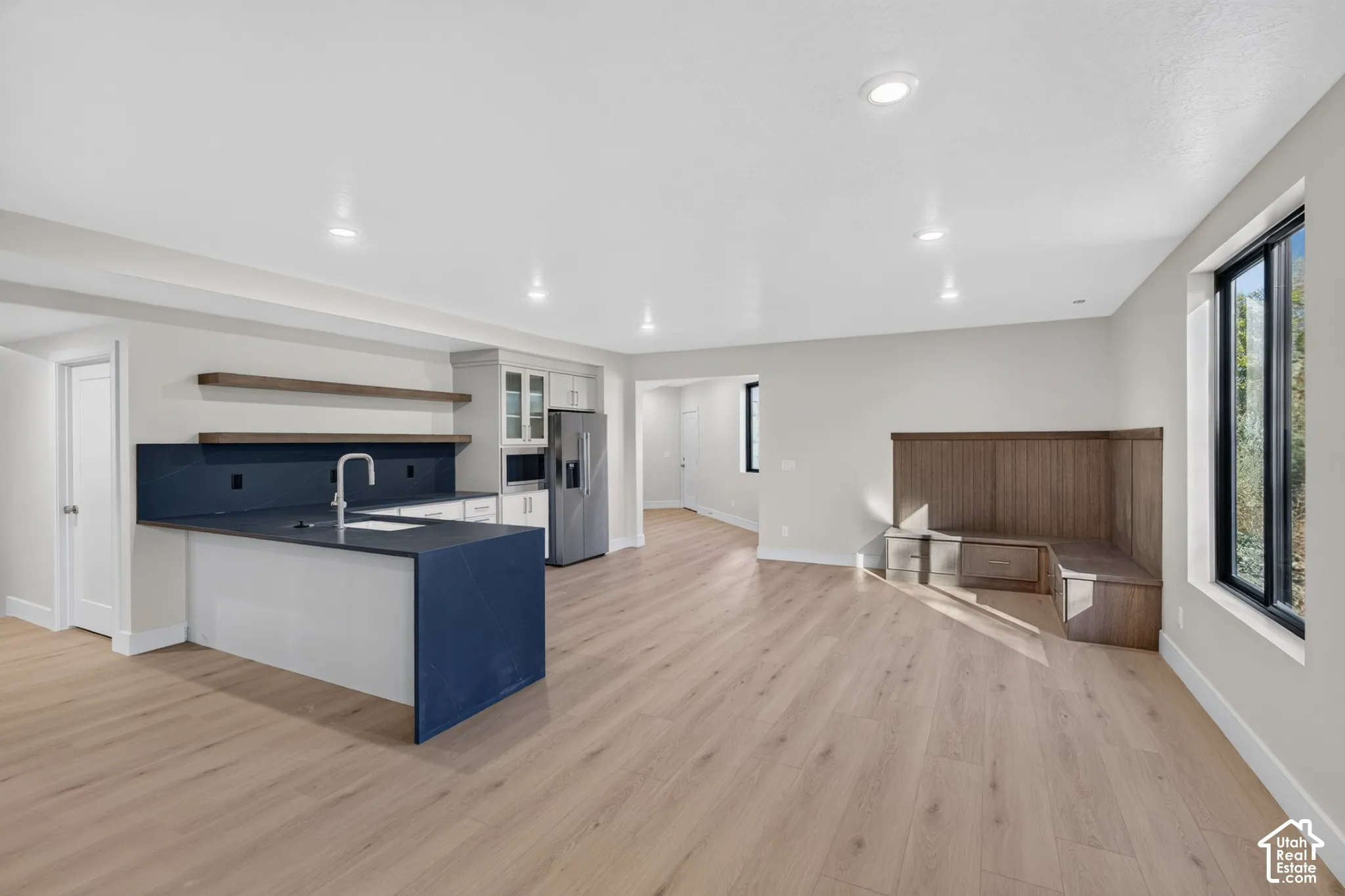 Open-concept kitchen featuring wood-finish flooring, a matte dark countertop with matching backsplash, floating wall shelving, and integrated wood-paneled bench seating