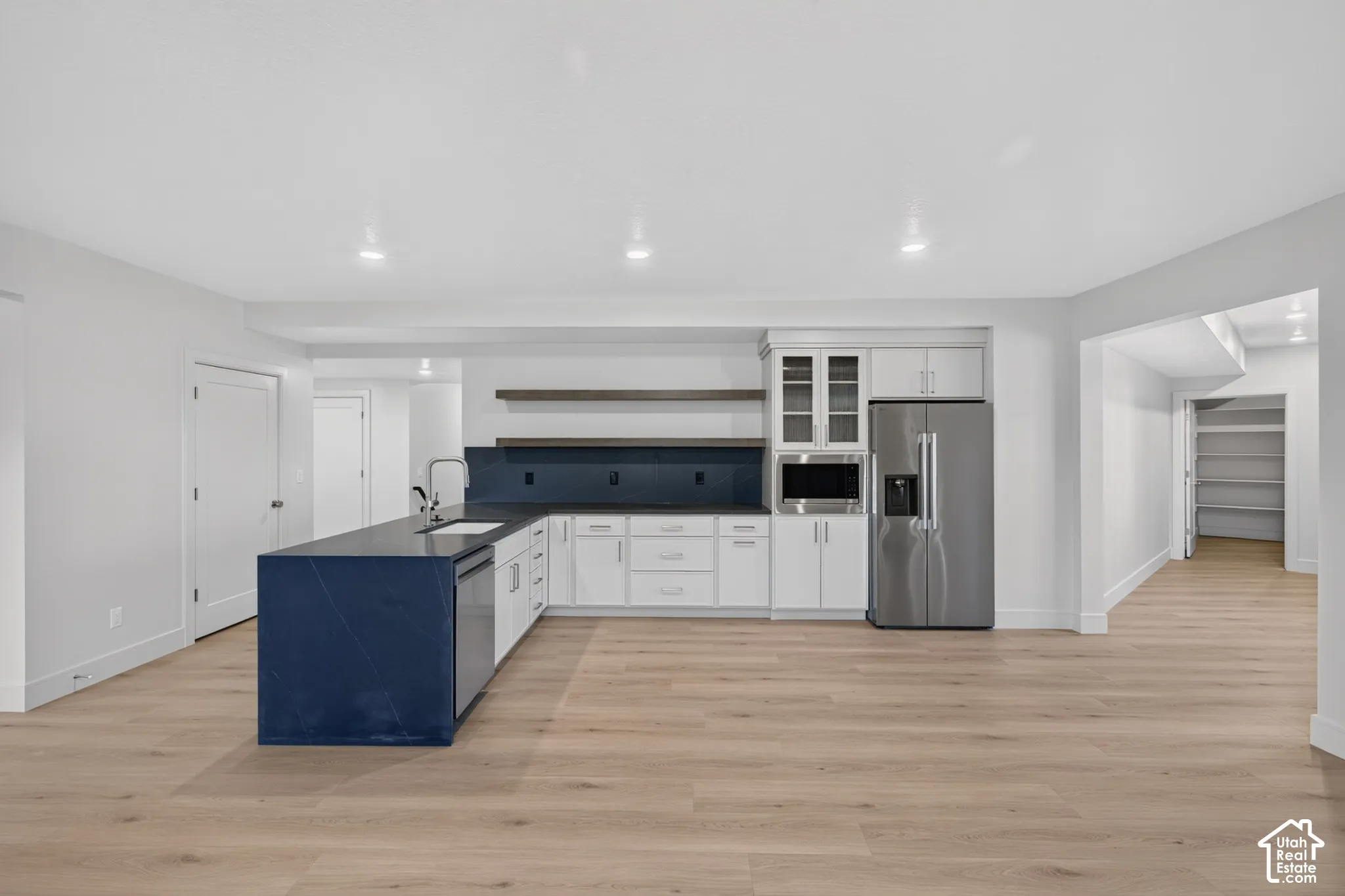 Kitchen featuring white cabinetry, a dark solid-surface island with an undermount sink, stainless steel appliances, open wood shelving, and wood-finish flooring