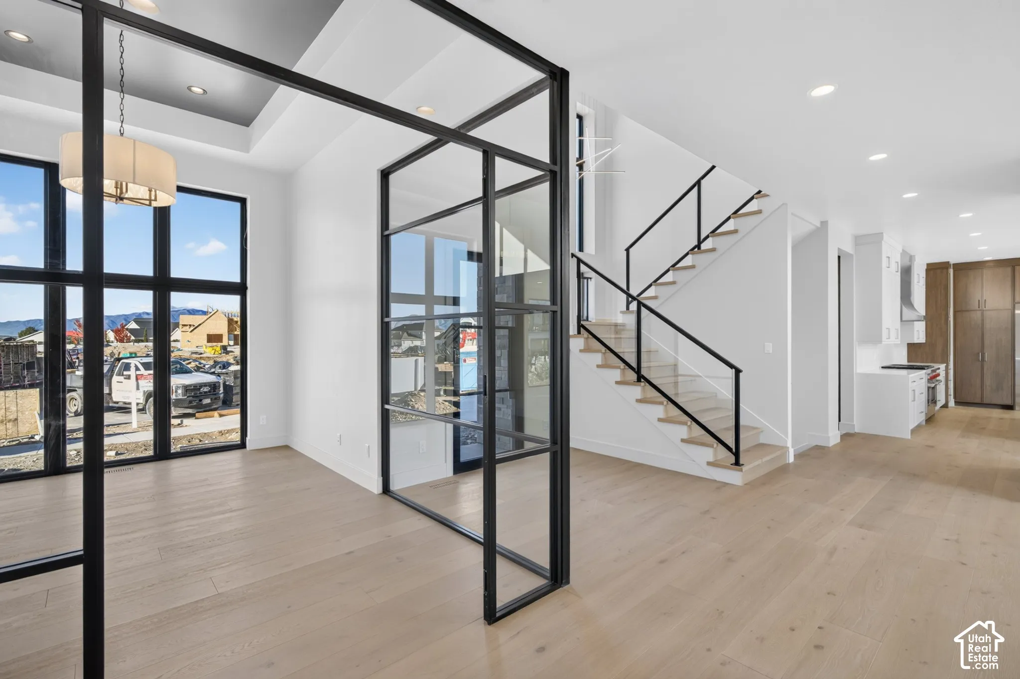 Open-concept interior featuring floor-to-ceiling black metal-framed glass partition walls, wood-finish flooring, tray ceiling with recessed lighting, and a staircase with matte black metal railings and wood treads