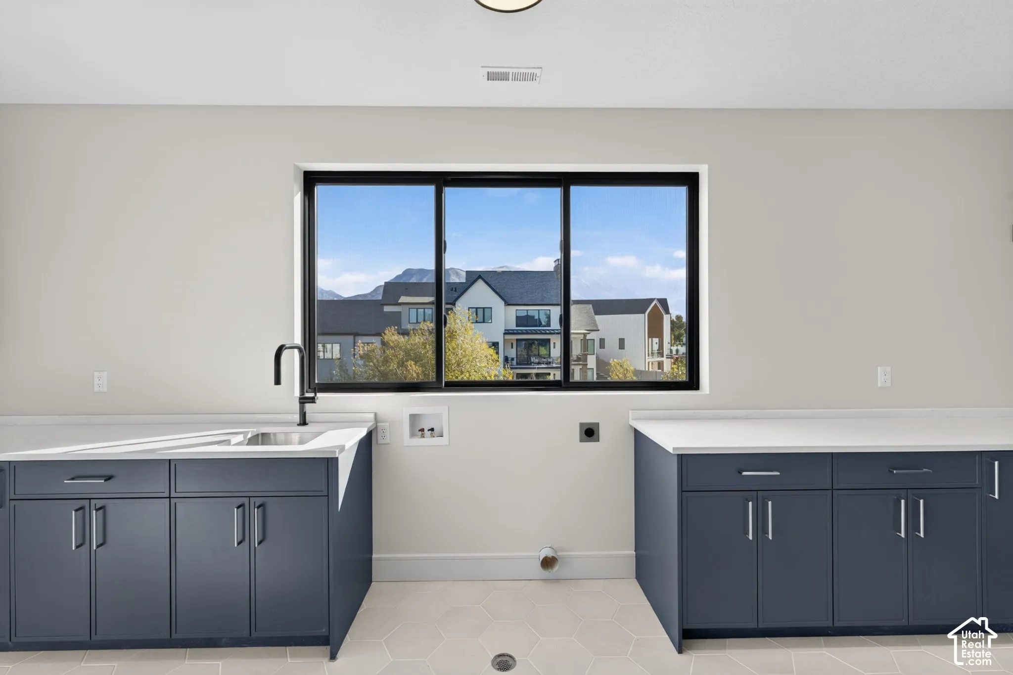 Dark-toned cabinetry featuring chrome hardware, white solid-surface countertops, a stainless steel sink with a matte black faucet, and hexagon-patterned floor tile