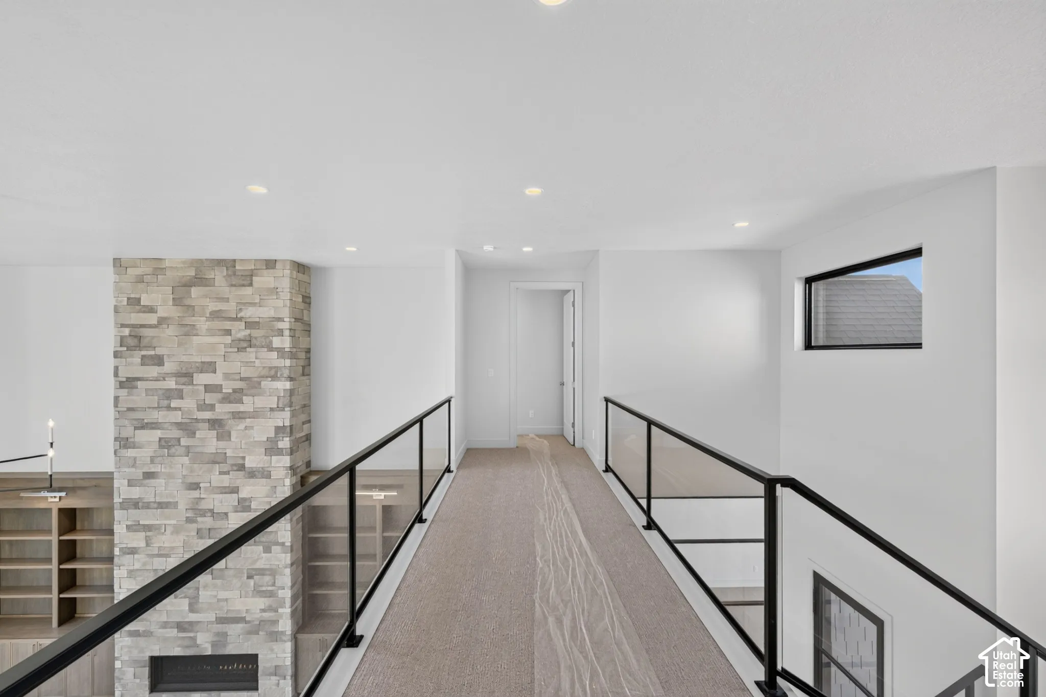 Elevated walkway featuring a floor-to-ceiling stone fireplace, sleek glass railing with matte black framing, neutral carpeting, recessed lighting, and a high-contrast rectangular window