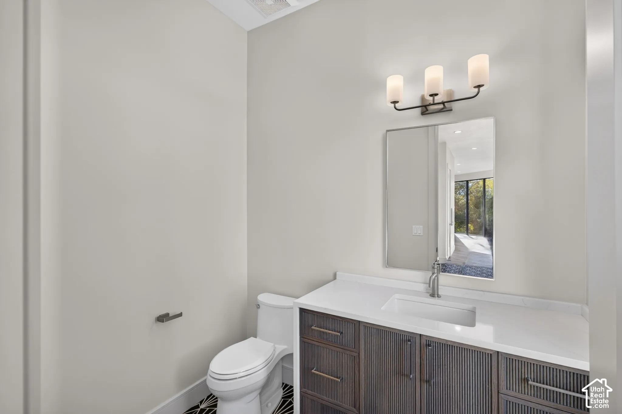 Bathroom vanity featuring textured dark-finish cabinetry, a white countertop with an integrated rectangular sink, a three-light wall-mounted fixture, and a vertical wall mirror