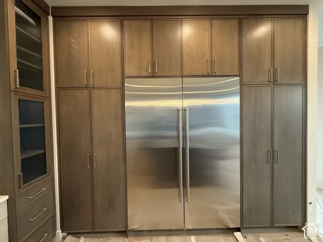 Floor-to-ceiling wood cabinetry featuring integrated paneling, stainless steel double-door refrigerator, and glass-front display shelving with matte metallic hardware