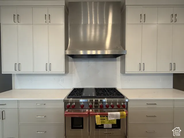 Stainless steel dual-fuel range featuring a heavy-duty professional-grade vent hood, light-toned shaker-style cabinetry with elongated hardware, and solid-surface countertop with coordinating full-height backsplash