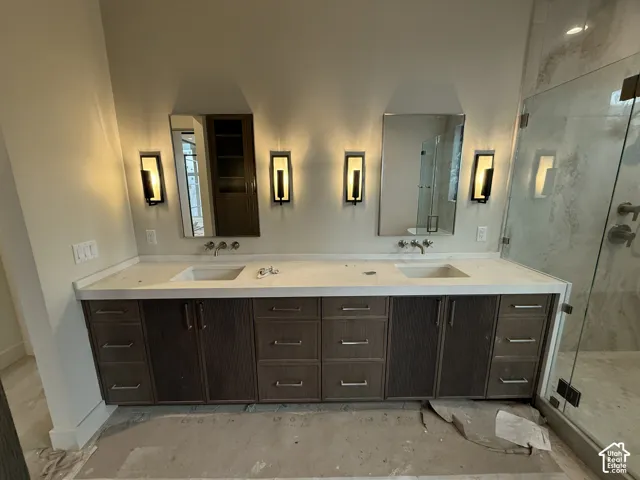 Dual-sink vanity with a solid surface countertop, dark wood-finish cabinetry, rectangular wall-mounted light fixtures, and a glass-enclosed shower