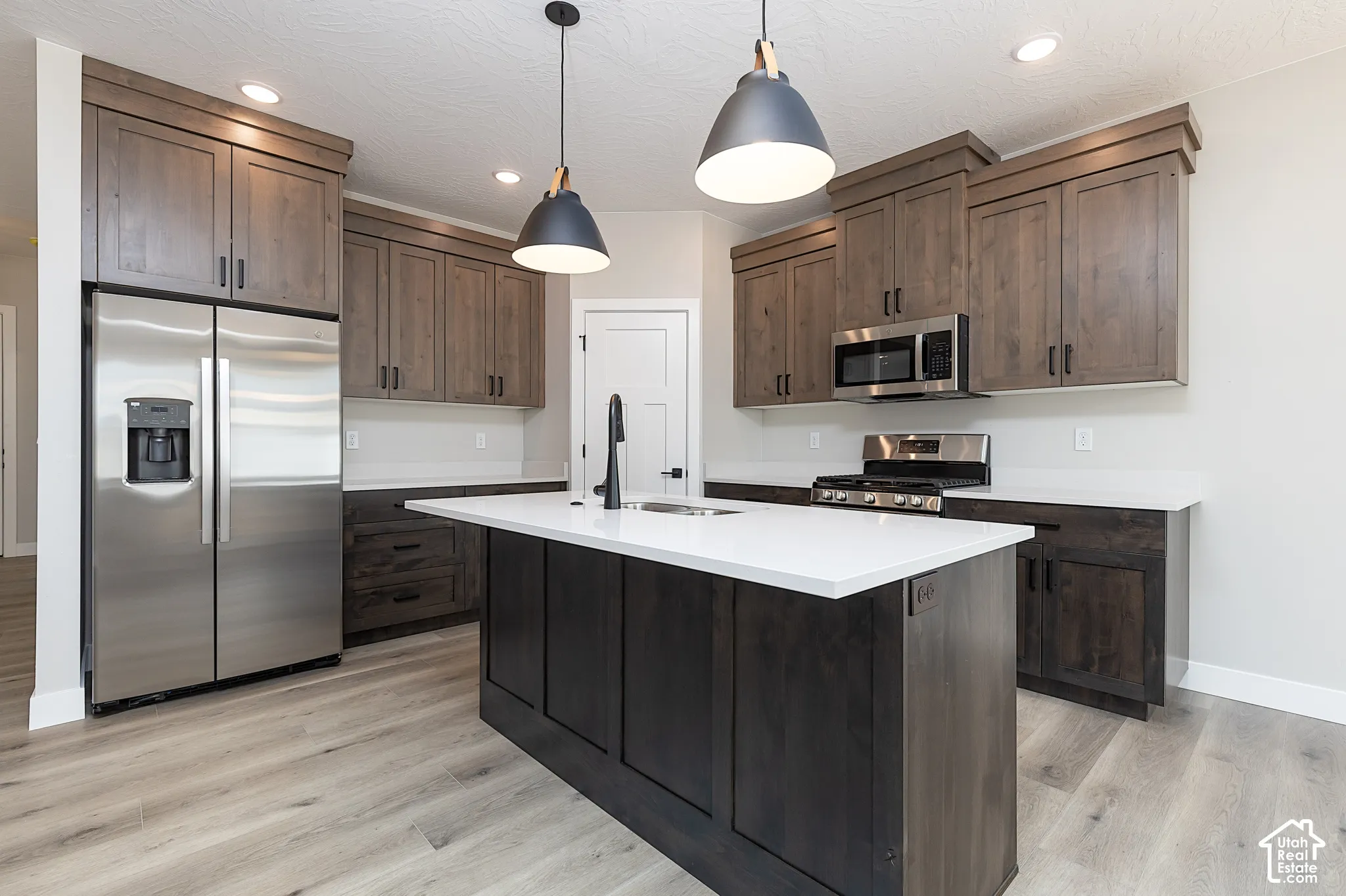 Kitchen featuring stained wood cabinetry, quartz countertops, a central island with an undermount sink, stainless steel appliances, and dual pendant lighting