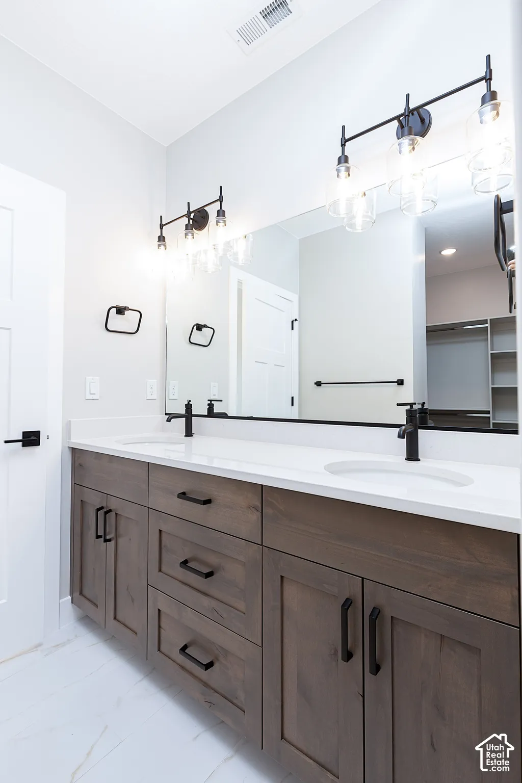 Dual-sink vanity featuring stained wood cabinetry, white stone-composite countertop, matte black hardware, and two multi-light vanity fixtures