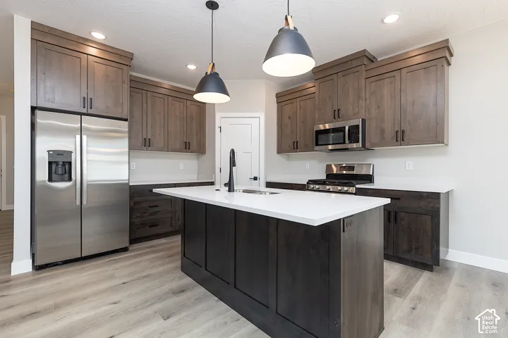 Kitchen featuring stained wood cabinetry, quartz countertops, a central island with an undermount sink, stainless steel appliances, and dual pendant lighting