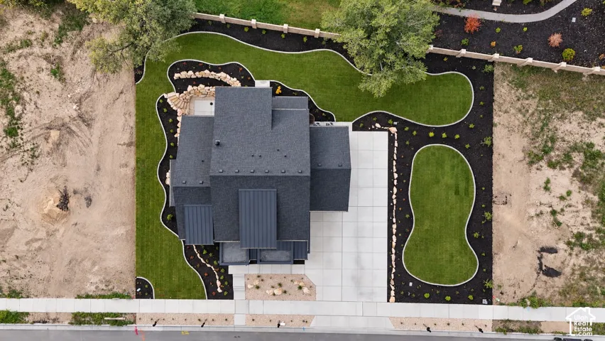 Aerial view of property featuring a multi-tonal shingle roof, concrete driveway, landscaped lawn with curved borders, dark mulch bedding, and rock accent features