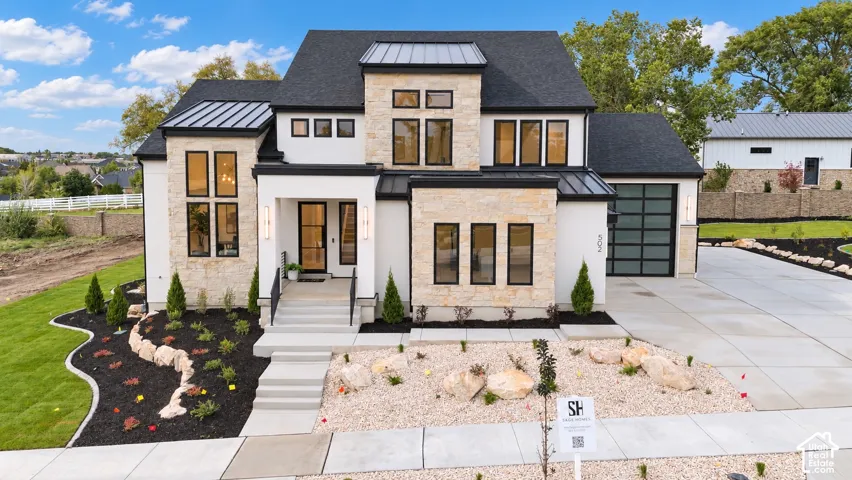 Modern exterior featuring a light-toned stone facade with smooth stucco accents, high-contrast dark window trim, a multi-gabled roof with metal roof accents, and a glass-paneled garage door