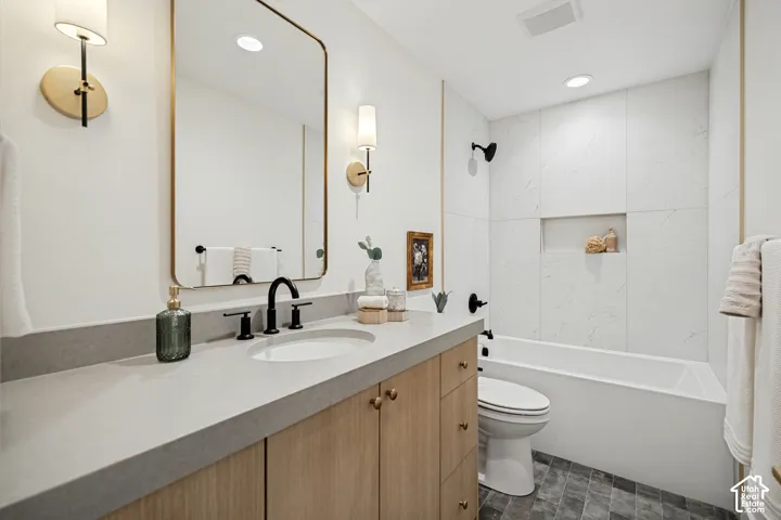 Bathroom featuring a wood-finish vanity with a stone-composite countertop, wall-mounted brass light fixtures, a rectangular mirror, a shower-tub combination with large-format wall tile and built-in niche, and dark-toned floor tiling
