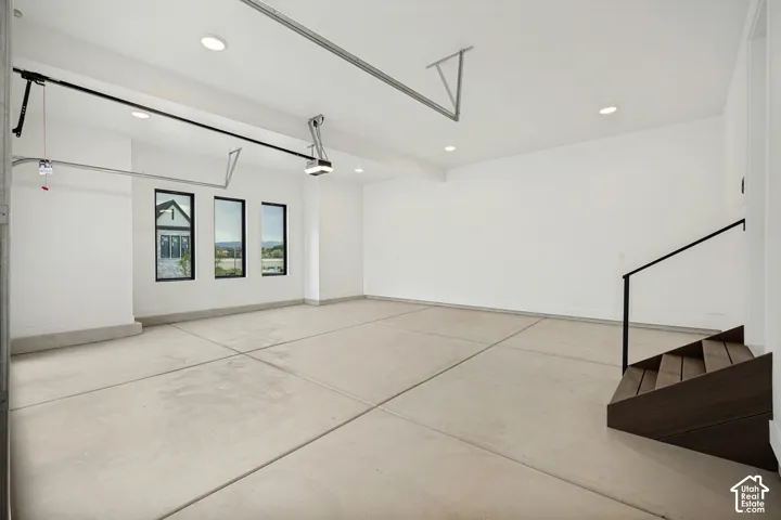 Finished garage interior featuring smooth white walls, recessed ceiling lighting, three black-framed windows, and a wood-tread staircase with a black metal railing
