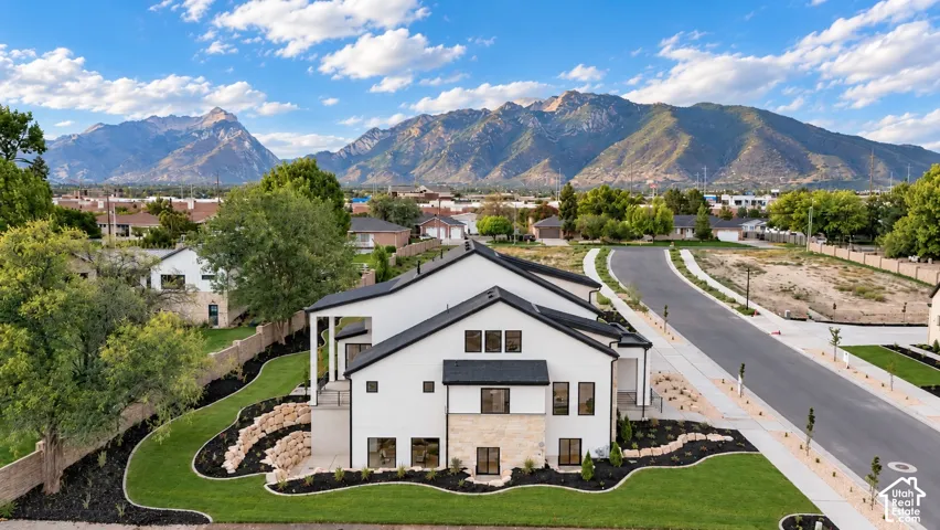 Two-story exterior featuring white stucco and stone veneer facade, steep-pitched metal roof, covered balcony, tiered rock garden landscaping, and prominent mountain backdrop