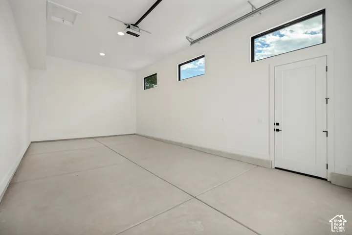 Finished garage interior featuring smooth concrete flooring, high-contrast windows, crisp white walls, recessed ceiling lighting, and a neutral-toned man door