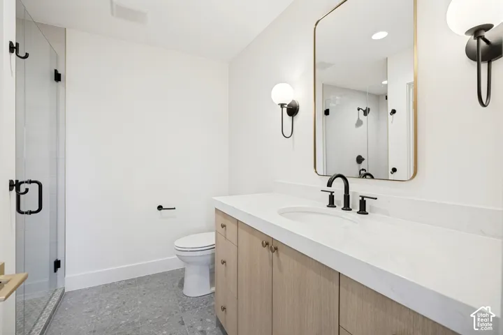 Bathroom featuring a light wood-finish vanity with a white countertop, wall-mounted globe lighting, a rectangular mirror with gold-tone frame, glass-enclosed shower, and grey stone-pattern flooring