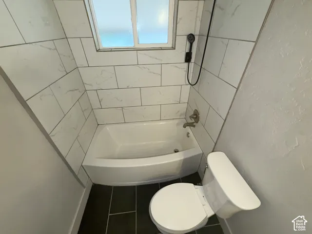 White subway tile shower surround, shower/tub combination, frosted window, black floor tiles, and a white commode