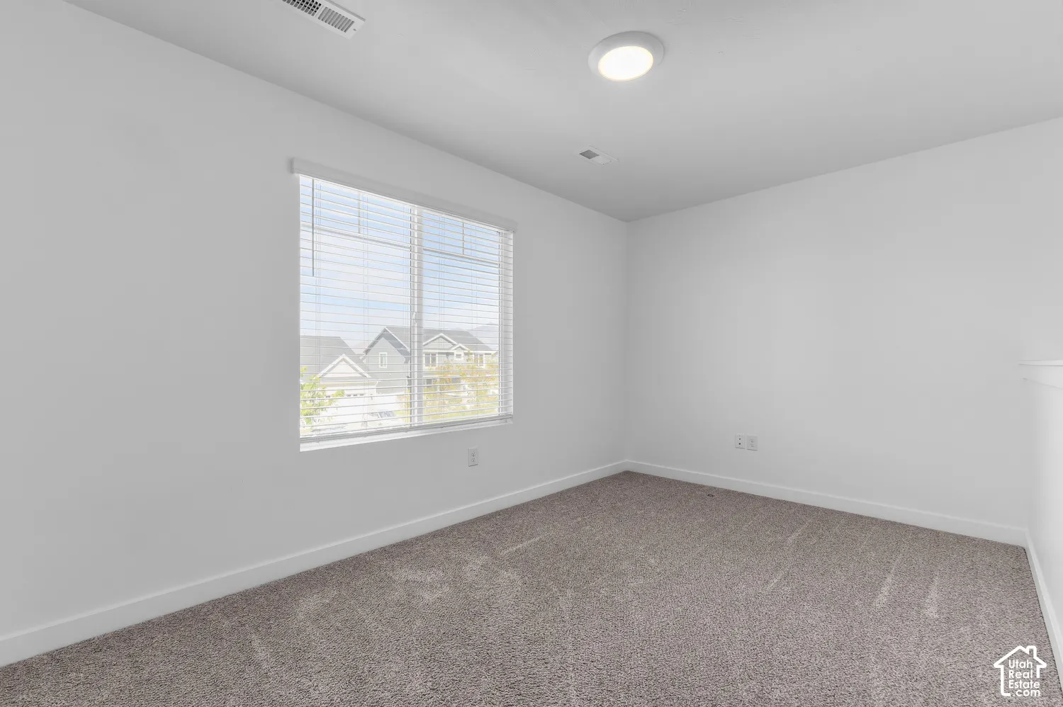 Neutral-toned wall-to-wall carpeting, crisp white walls, flush-mount ceiling lighting, and large window with horizontal blinds
