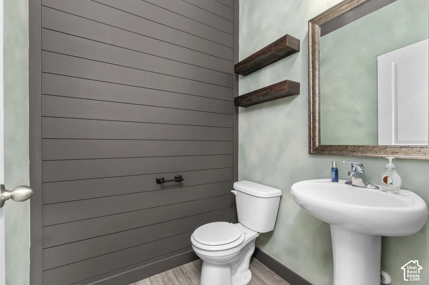 Dark-toned horizontal shiplap accent wall, two floating wood-finish shelves, pedestal sink with chrome faucet, and wood-finish flooring