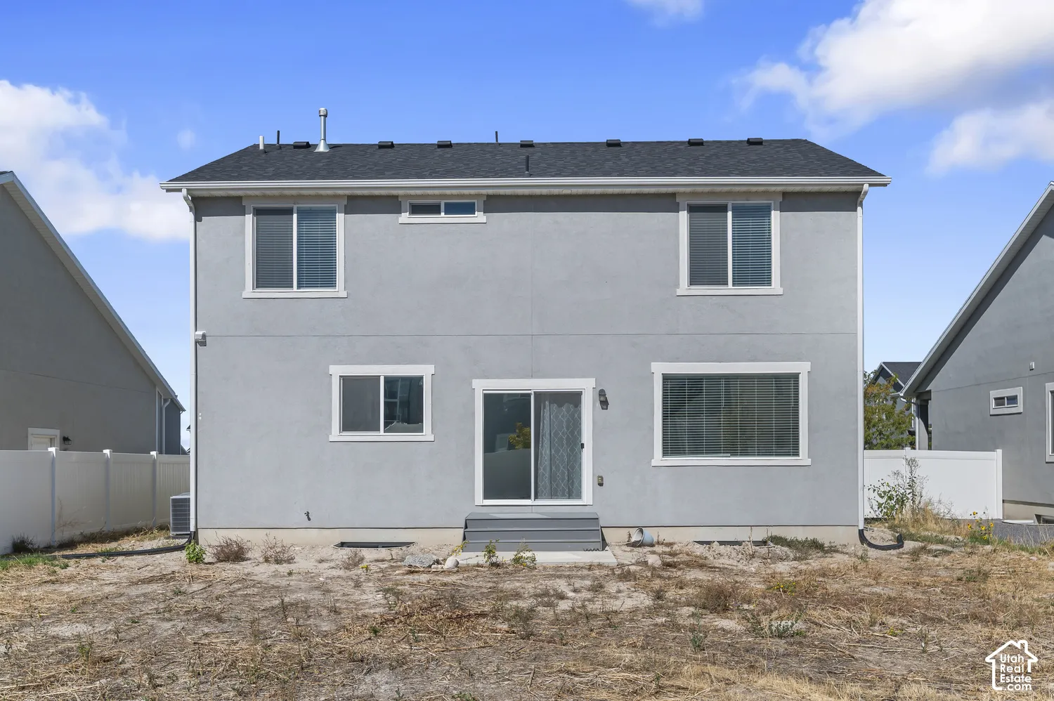 Two-story exterior featuring light gray stucco siding, asphalt shingle roofing, sliding glass door, and white vinyl privacy fencing