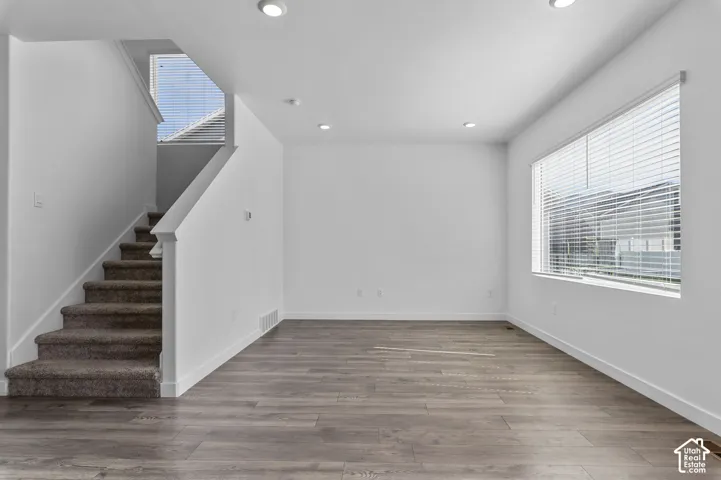 Neutral-toned interior space featuring wood-finish flooring, recessed lighting, expansive windows with horizontal blinds, and a staircase with carpeted treads