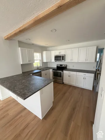 Kitchen featuring white cabinetry, dark countertops, wood-finish flooring, stainless steel appliances, and an exposed ceiling beam