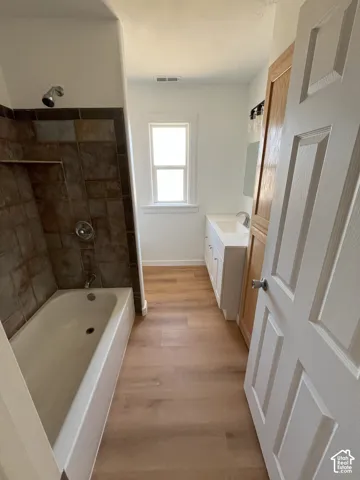 Bathroom featuring wood-finish flooring, a white vanity with an integrated sink, a tiled bathtub surround, and a built-in wooden storage cabinet