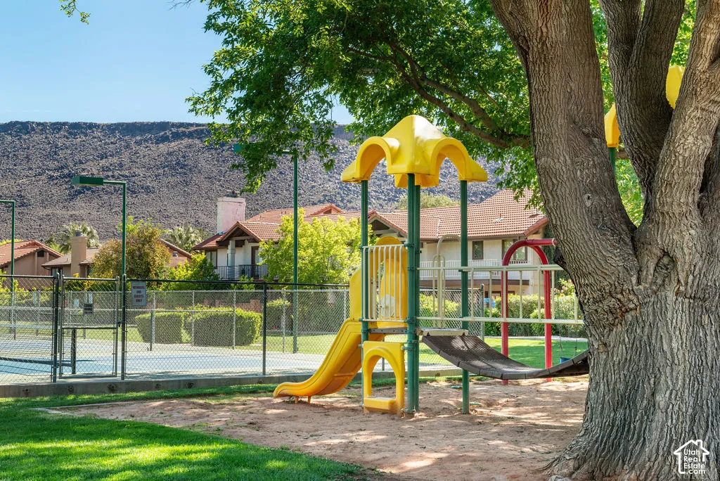 Playground equipment with slides and climbing features, shaded by mature trees, bordering a fenced tennis court