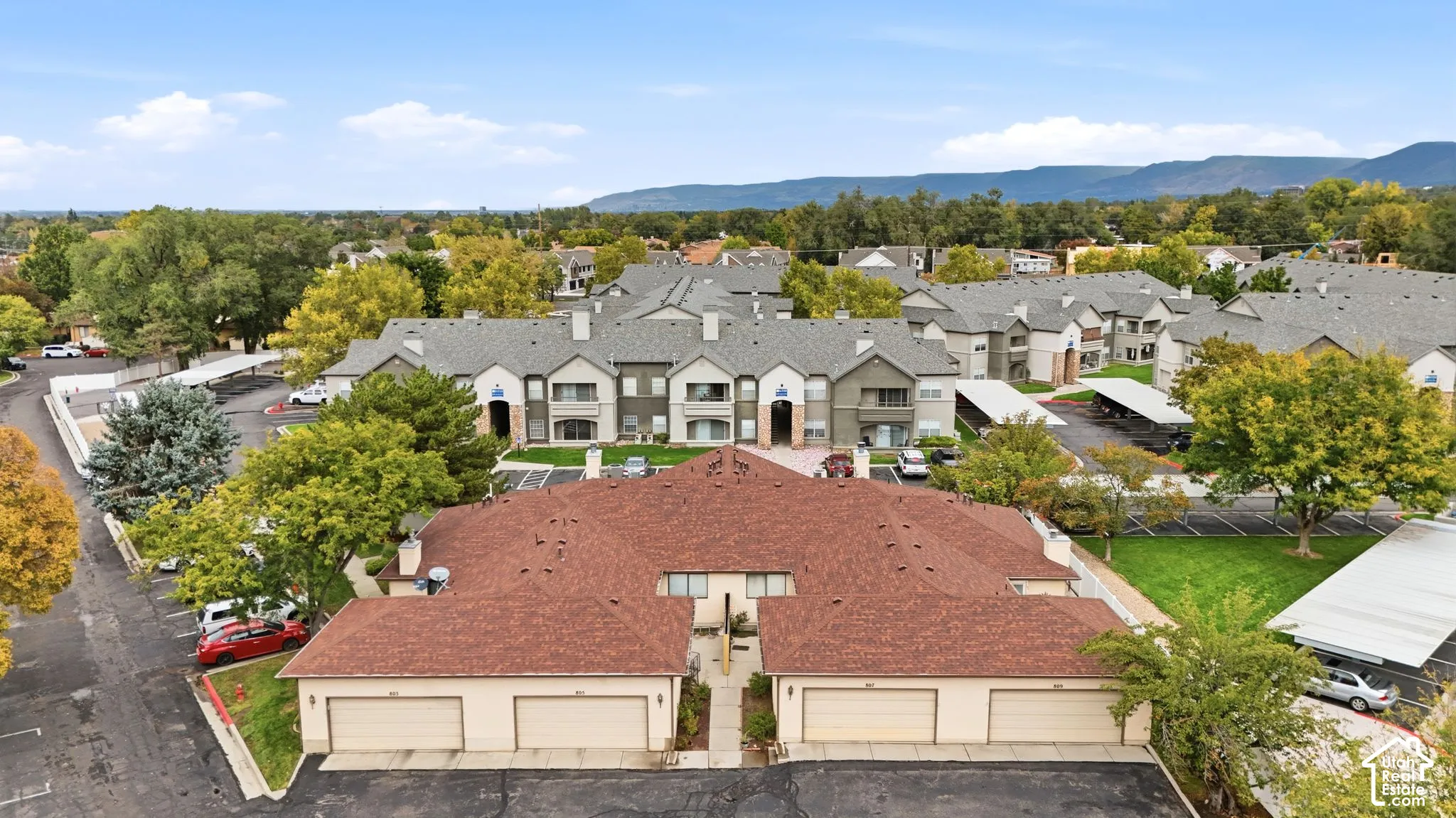 Multi-building residential complex featuring shingled hip roofs, stucco siding, detached garage structures, covered parking carports, and mature trees against a mountain backdrop