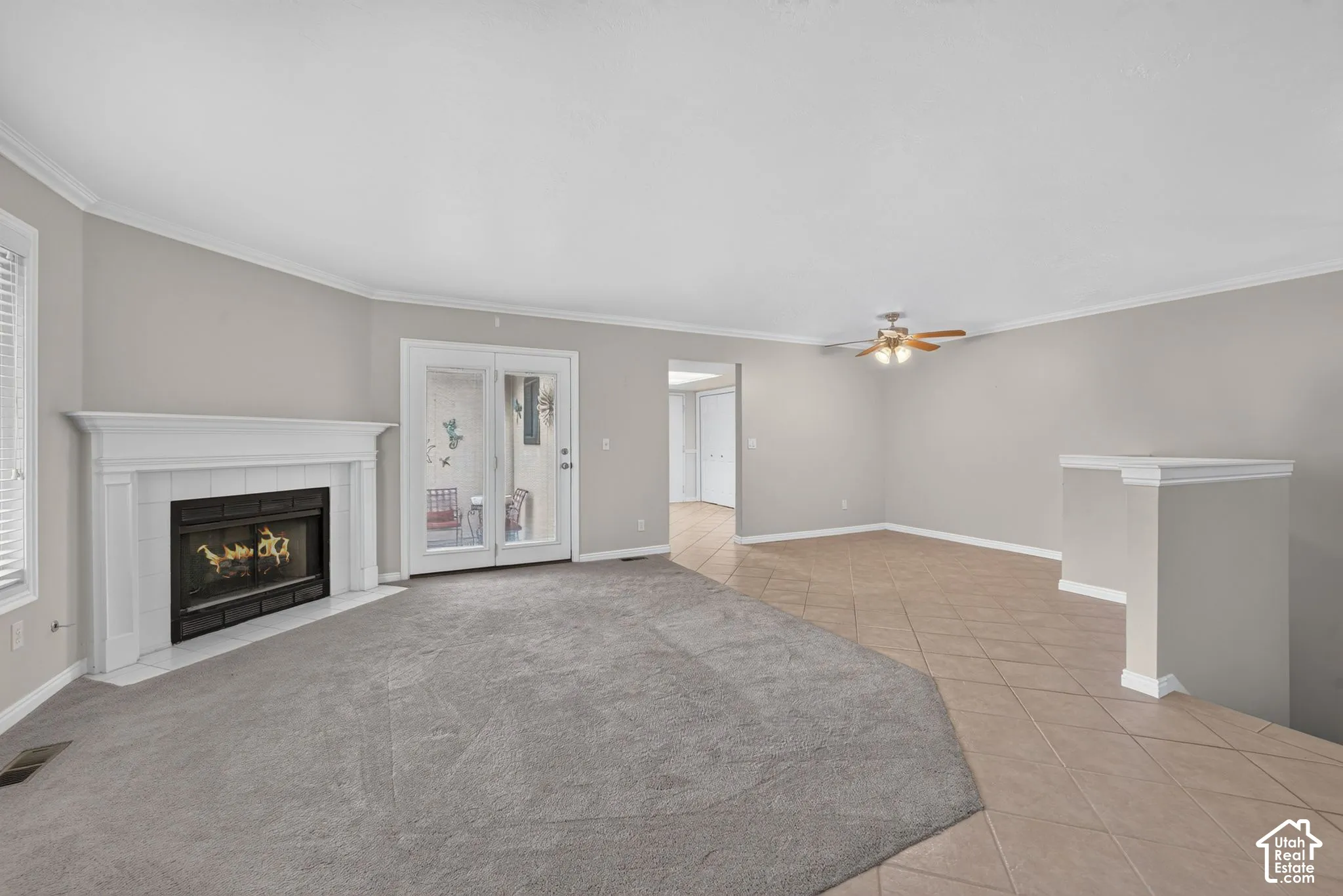 Fireplace with a classic white mantel, neutral-toned wall-to-wall carpeting, diagonal tile flooring, glass-paneled double doors, and a ceiling fan with integrated lighting