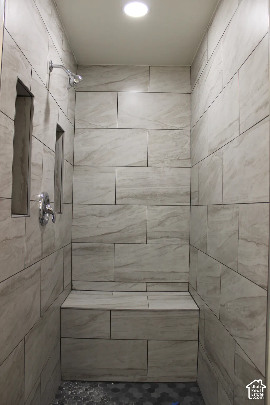 Walk-in shower featuring large-format neutral tile walls, an integrated tiled bench, dual recessed wall niches, and a contrasting hexagonal tile floor with polished chrome fixtures